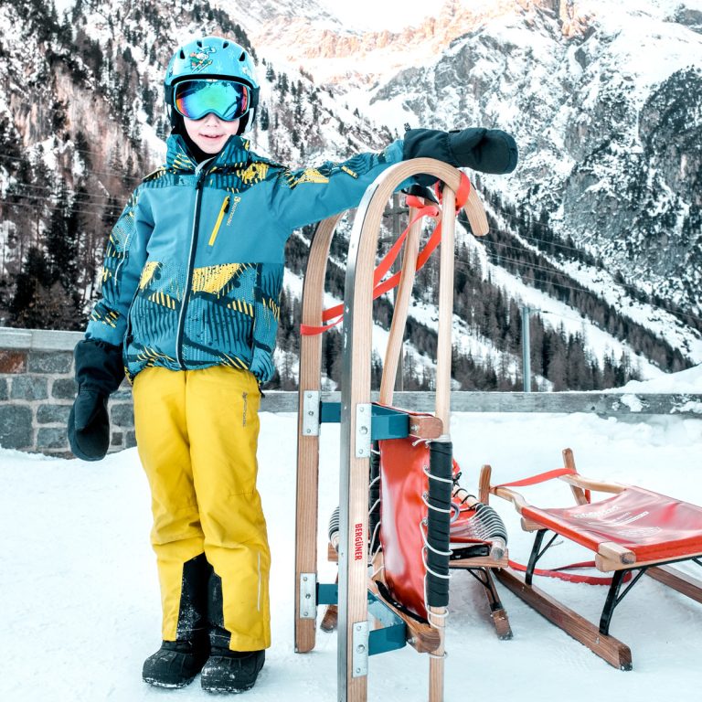 Junge mit Helm und Skibrille neben einem BERGÜNER Rodel mit roter Sitzbespannung vor Bergkulisse
