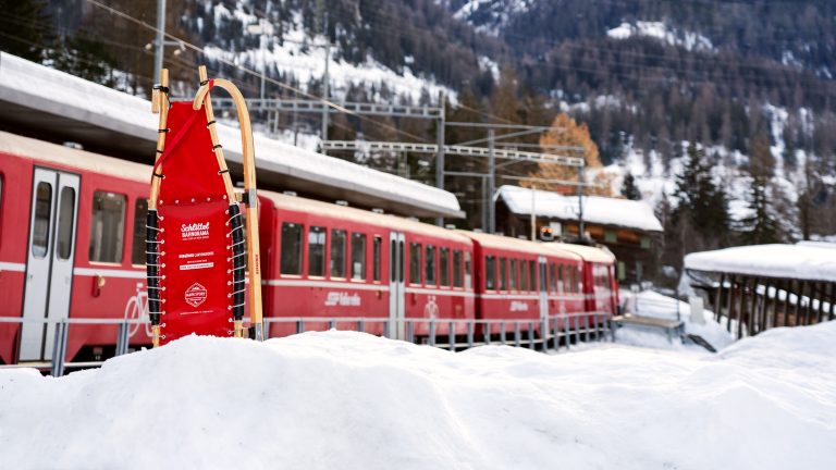 Roter Mietschlitten von Schlittel-Bahnorama im Schnee, dahinter ein roter Zug der Rhätischen Bahn am Bahnhof Bergün