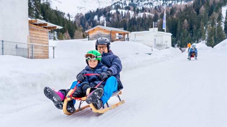Erwachsener und Kind mit Helmen fahren gemeinsam auf einem Holzschlitten die Schlittelbahn hinunter